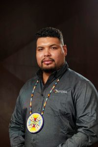 Indigenous man from Wisconsin with a short dark head of hair and facial hair wearing a long-sleeved gray shirt on a black background